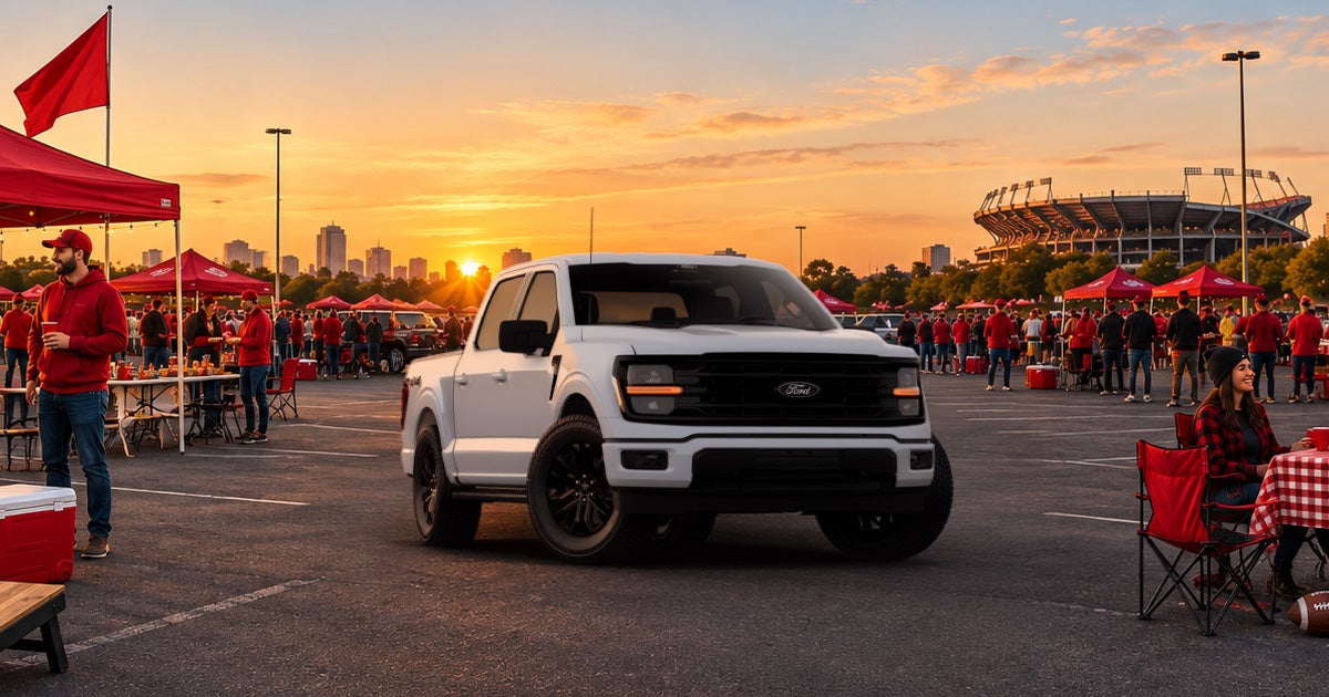 A white Ford F-150 pickup truck featuring a dark grille and black wheels parked at a tailgate in front of a stadium.